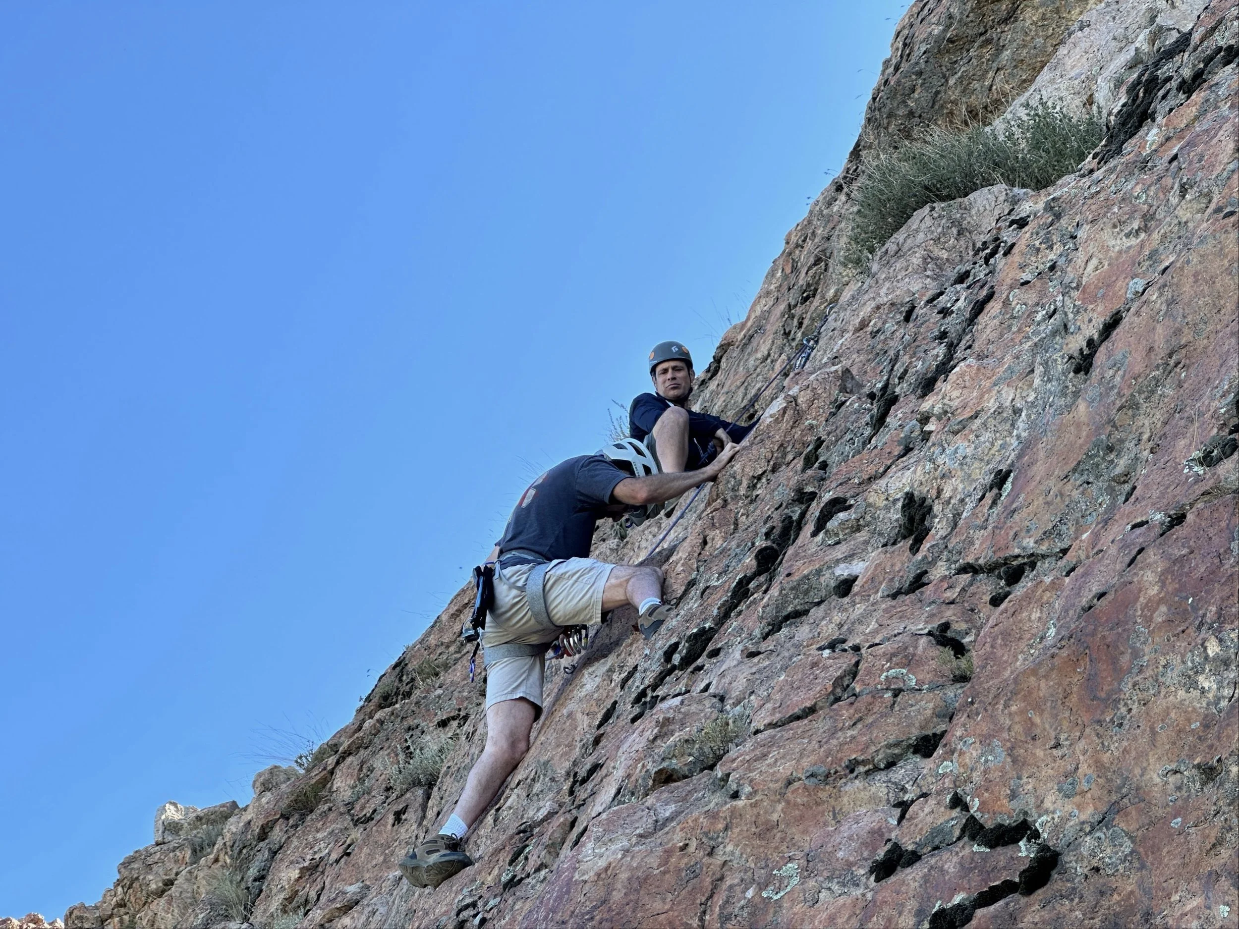 Two men rock climbing on a steep, rocky cliff under a clear blue sky.