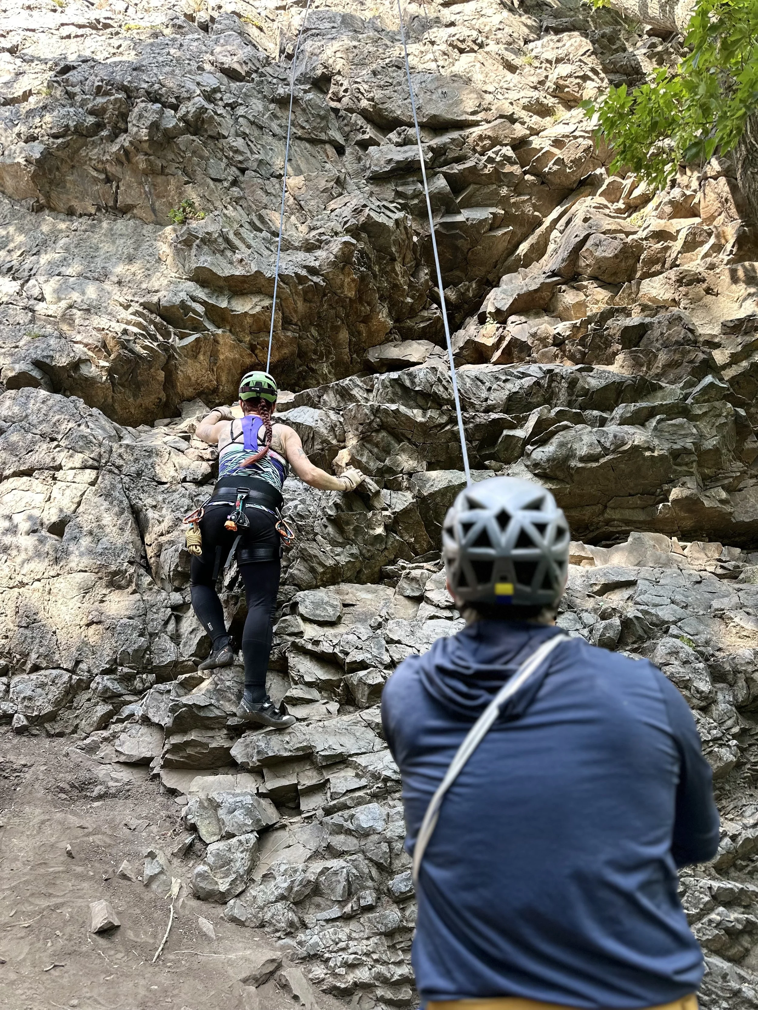 A person rock climbing on a rugged rock face while another person, wearing a helmet, watches nearby.