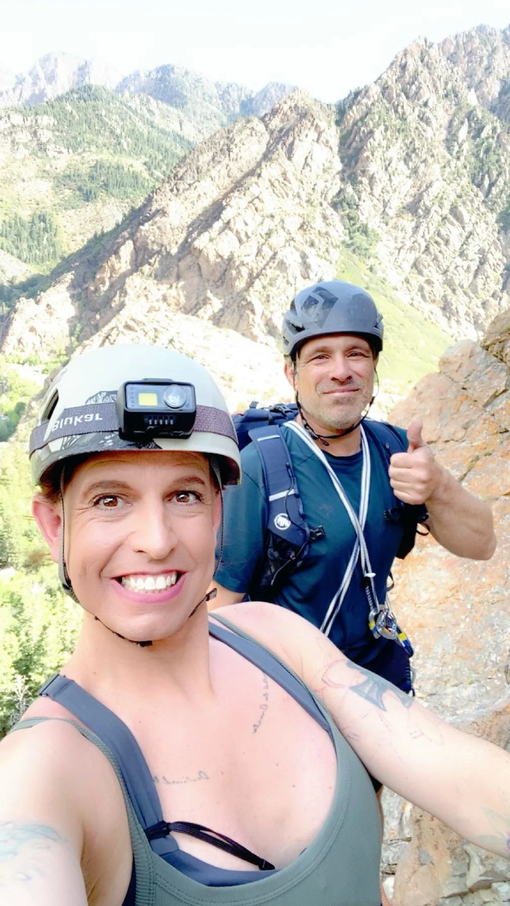 Two people wearing helmets and hiking gear taking a selfie on a mountain trail with rocky peaks and green forest in the background.