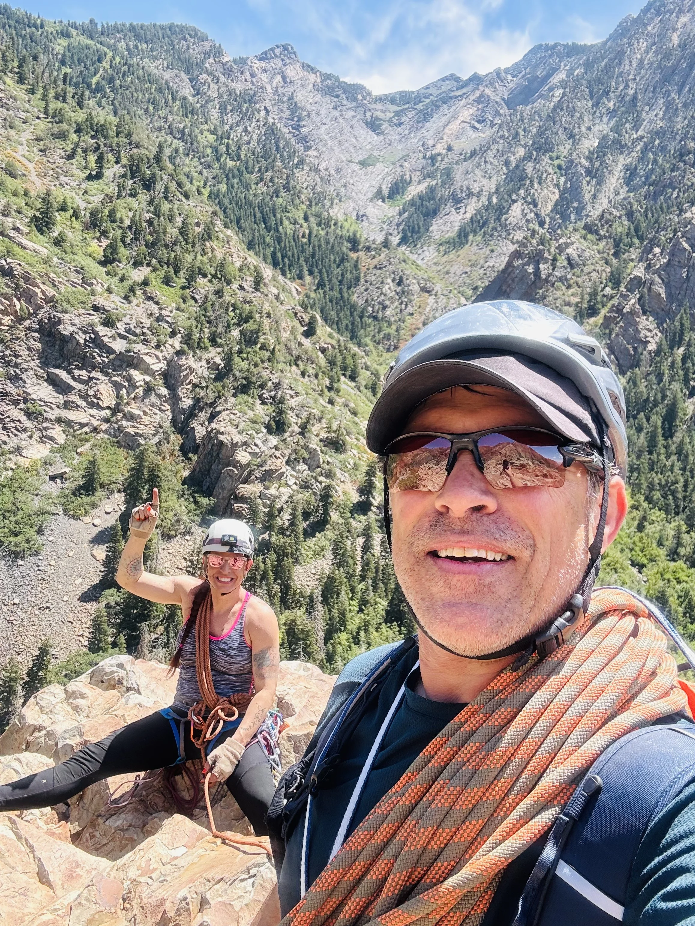 A man and woman in hiking gear on a rocky mountain ledge with a scenic mountain and forest background, both smiling and wearing helmets and sunglasses.