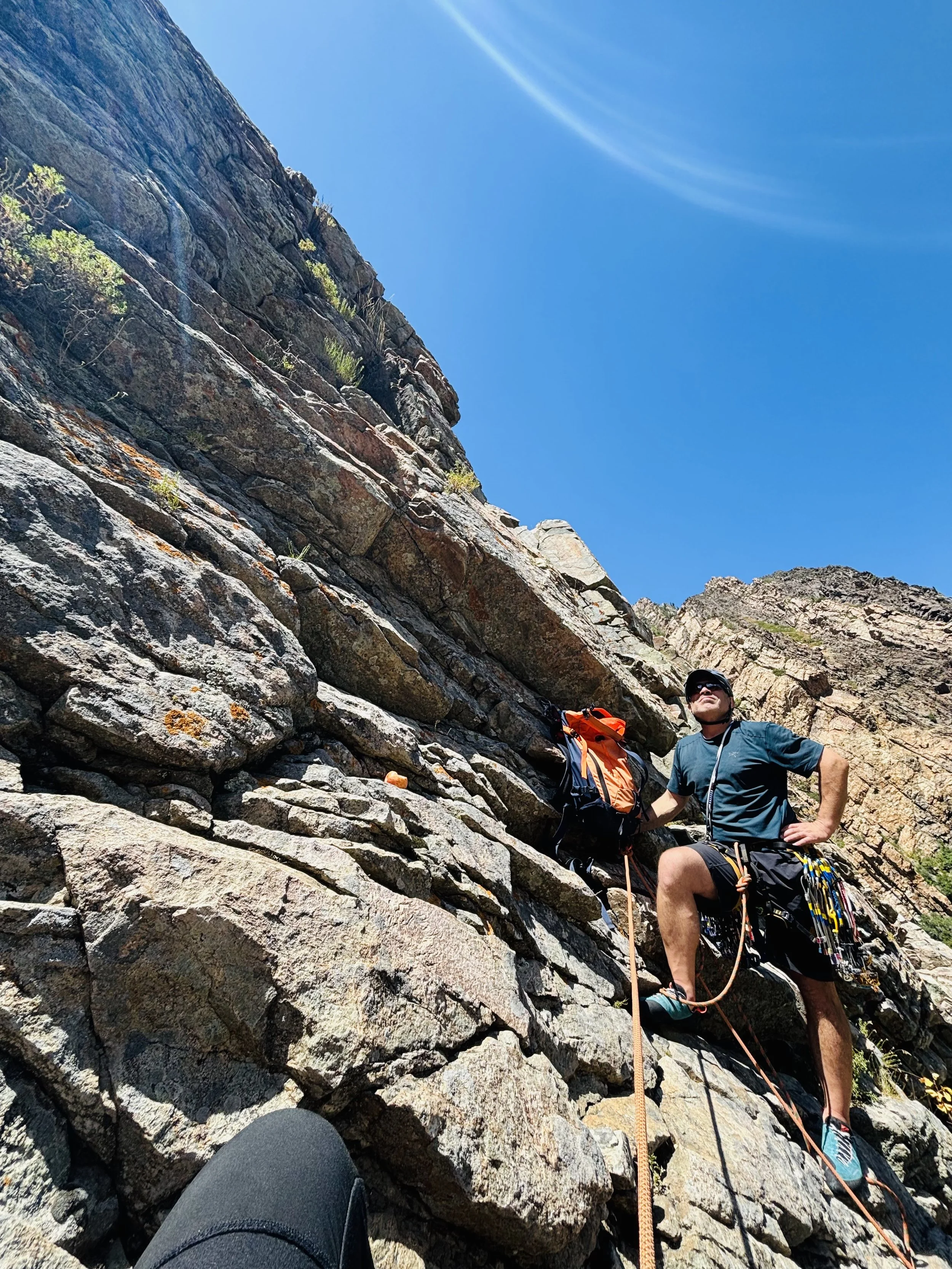 A person rock climbing on a steep, rocky mountain slope with climbing gear and a backpack, under a clear blue sky.