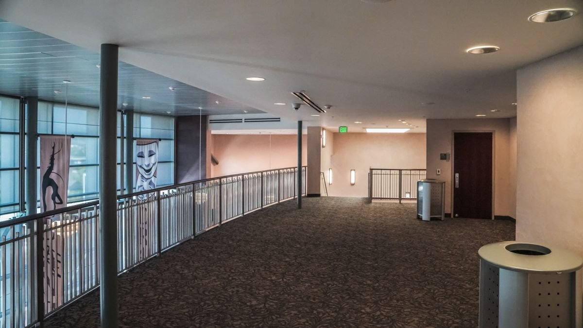 Interior view of a modern building corridor with large windows, decorative banners, and a railing, with a trash can and an elevator door.