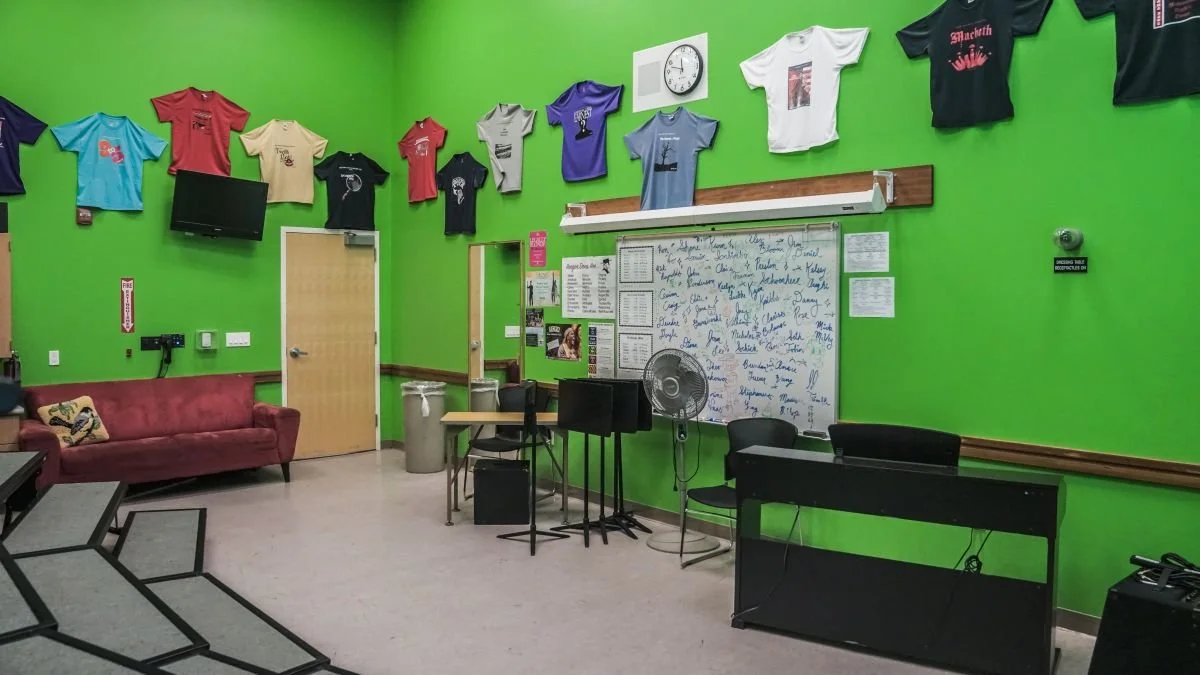 A vibrant green room decorated with various T-shirts on the wall, a small TV, a red sofa, a whiteboard with signatures or writing, and music or office equipment.