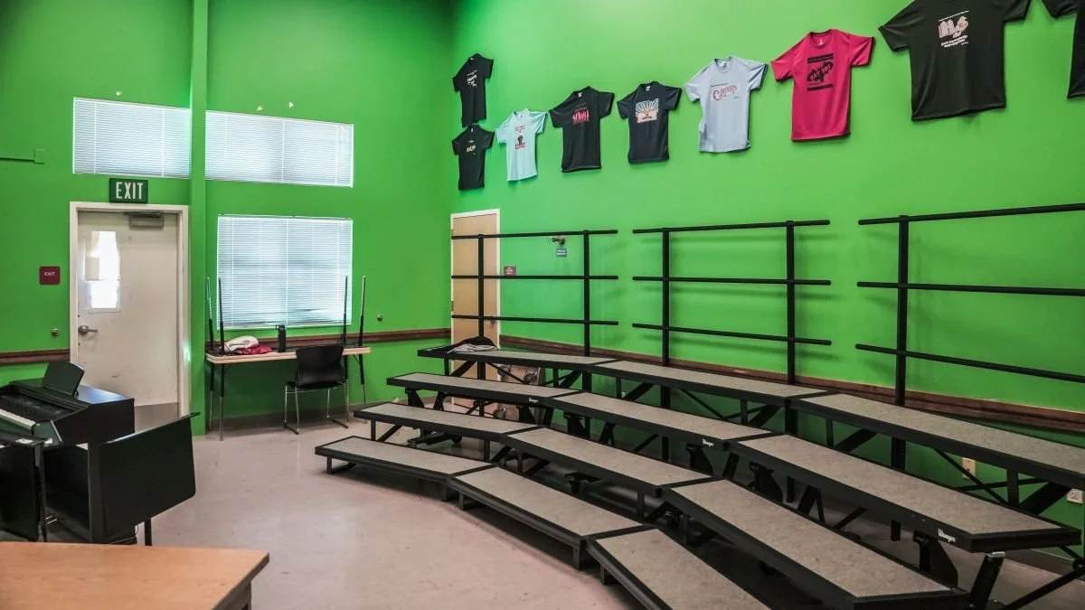 An empty performance or rehearsal room with green walls and tiered seating. T-shirts are hung on the wall, and there is a door, window blinds, a desk with a chair, and a piano in the room.