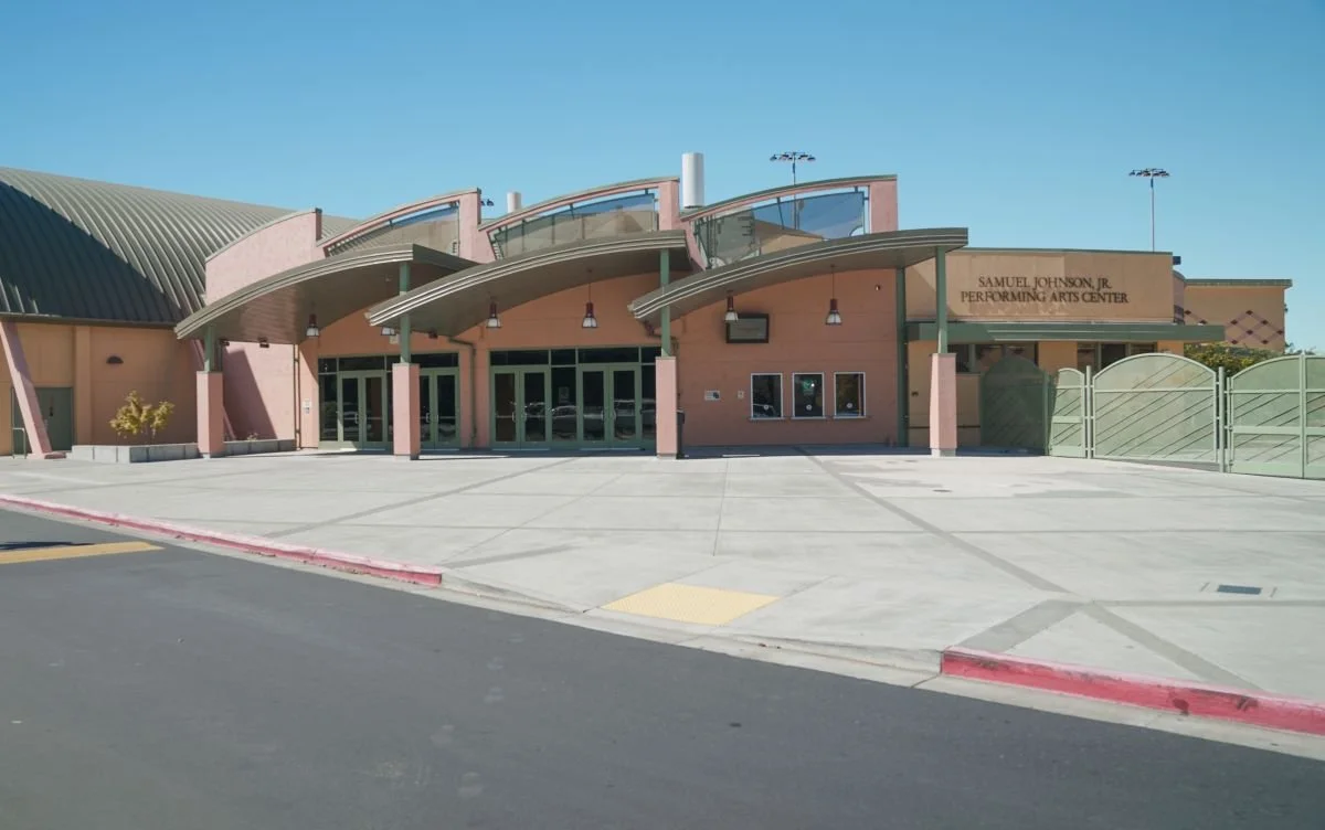Front view of the Samuel Johnson Jr. Performing Arts Center, a modern building with curved roof design, glass doors, and a paved parking area.