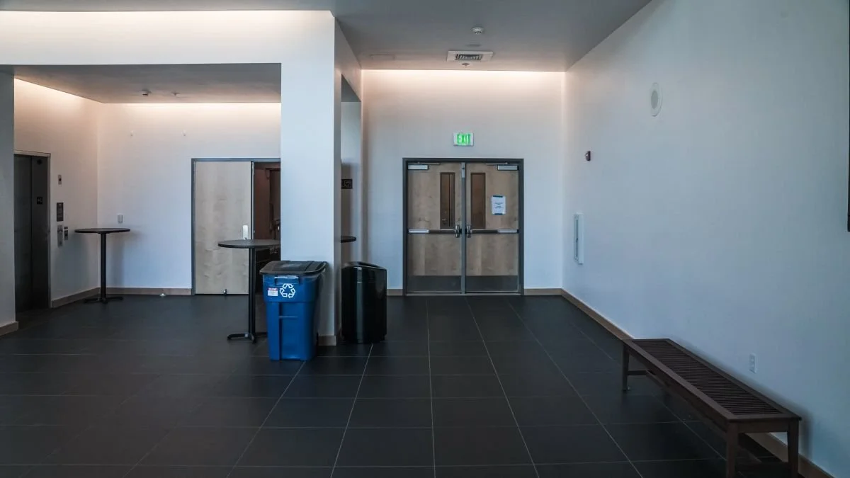 Empty room with black tiled floor, white walls, two blue recycling bins, and two closed wooden doors with an exit sign above one.