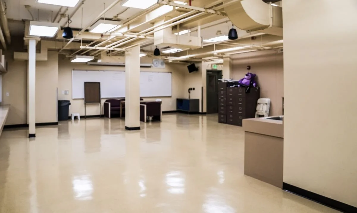 Empty classroom or activity room with beige walls and floor, white ceiling with exposed pipes, whiteboard, and furniture including cubbies, lockers, chairs, and a purple backpack.
