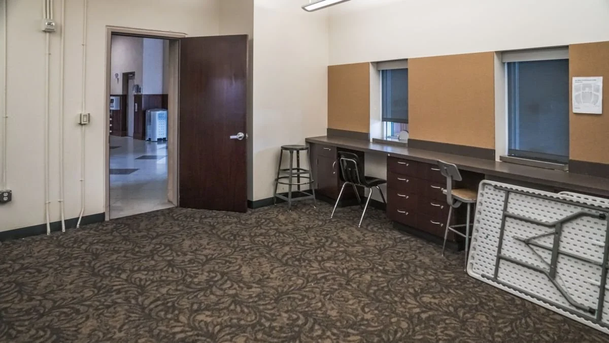 An empty room with a patterned carpet, a wooden door partially open, a long counter with three chairs, and a folding table leaning against the wall, with two small windows and plywood boards behind the counter.