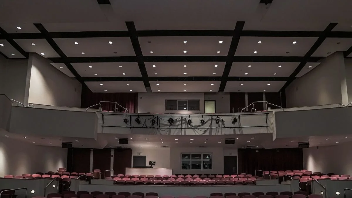 Empty auditorium with theater seats, stage, and balcony, illuminated ceiling lights, and sound equipment on the balcony railing.