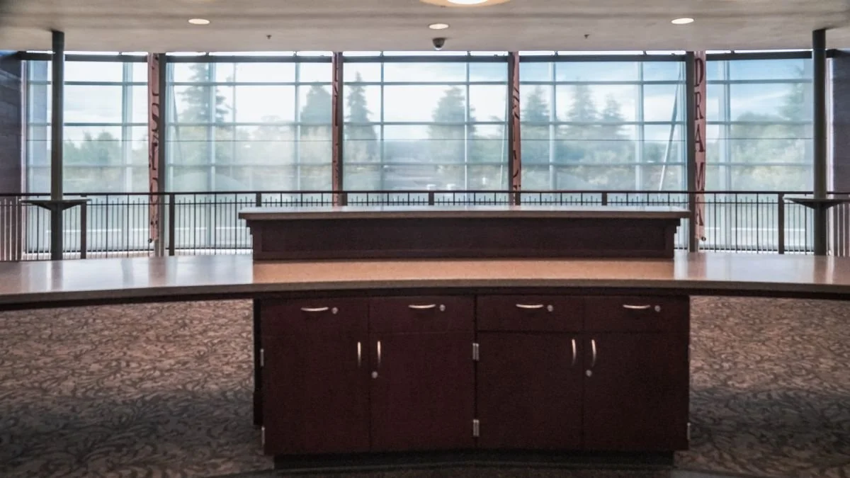 Empty indoor space with large windows, wooden counter in front, and carpeted floor.