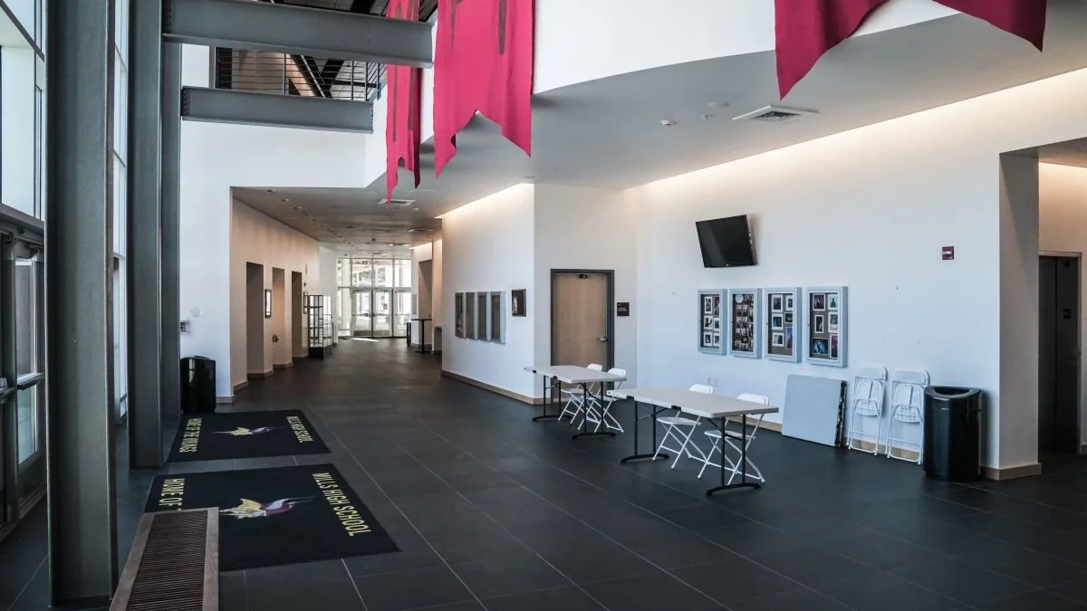 Modern interior of a building with white walls, black tiled floor, pink decorations hanging from the ceiling, and tables and chairs stacked against the wall.