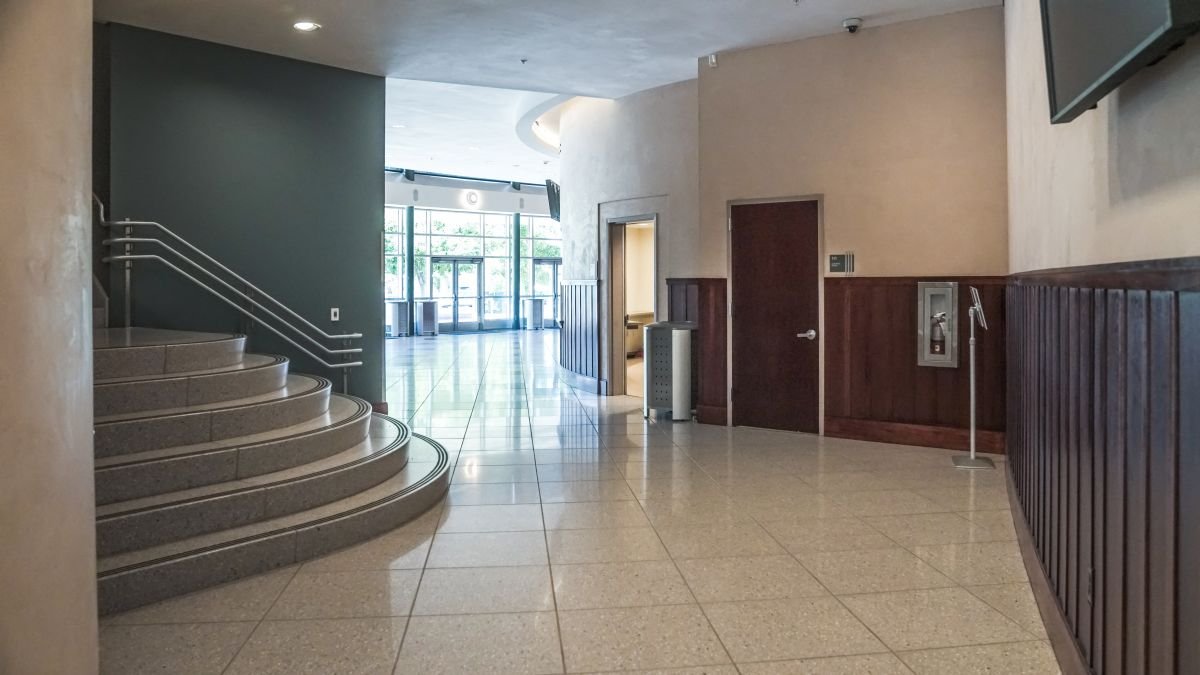 Empty lobby area with tiled floor, staircase on the left, and glass entrance doors at the back, with sunlight coming through.