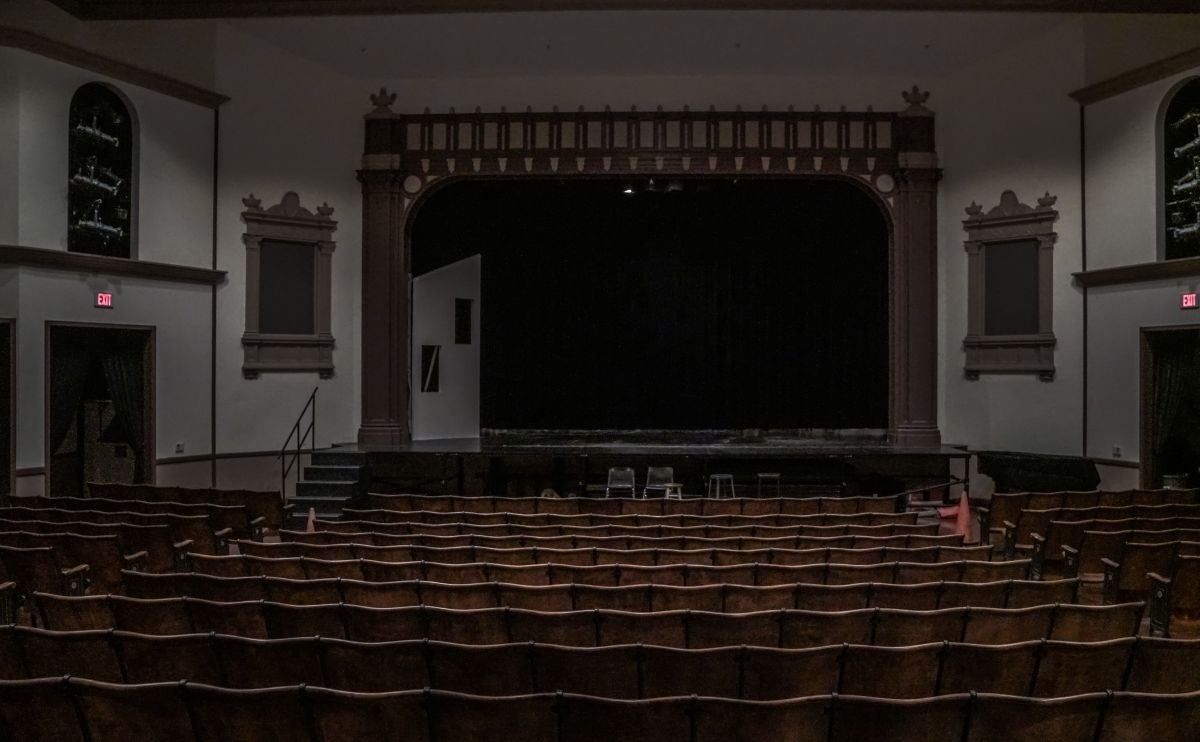 An empty theater with rows of wooden seats facing a stage with a black curtain, wooden trim, and an ornate proscenium arch.