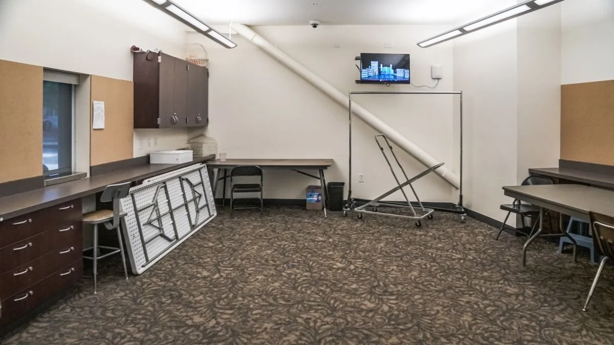 Empty room with tables and chairs, a TV on the wall, a window, and a large rolled-up whiteboard.