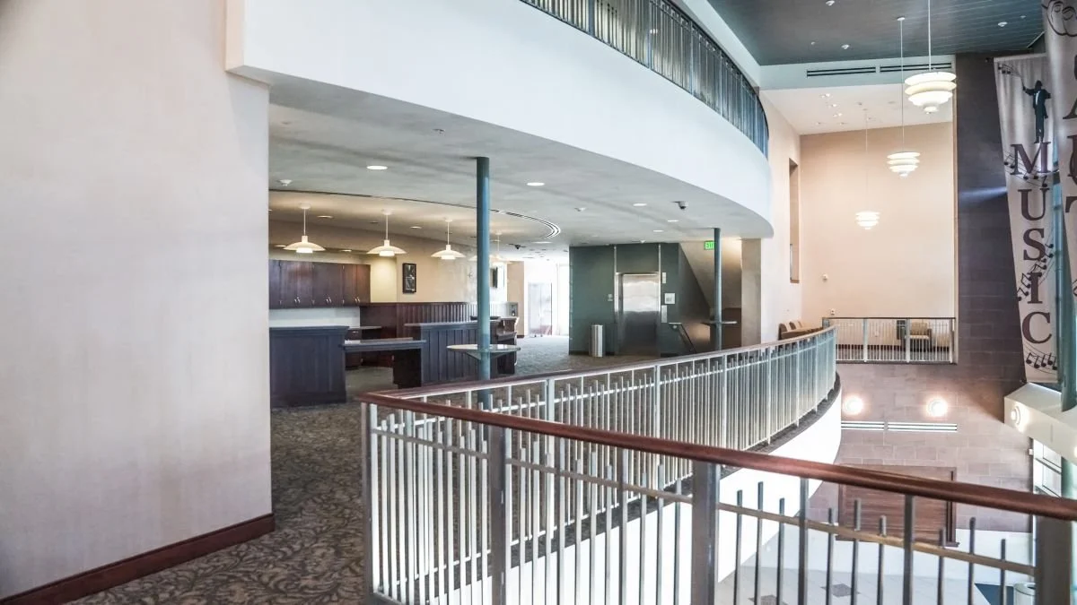 Interior view of a modern building with a curved balcony, railing, and multiple levels, featuring seating areas and hanging lights.