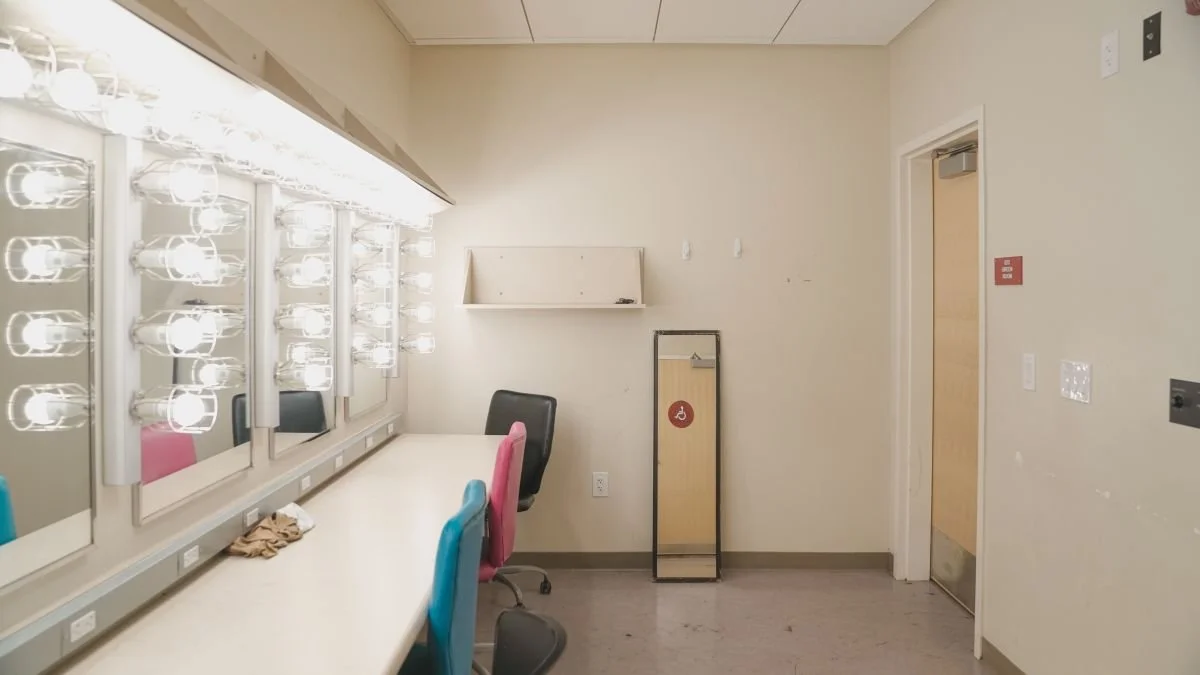 Empty dressing room with illuminated mirrors and chairs, including a wheelchair accessible sign on a mirror.