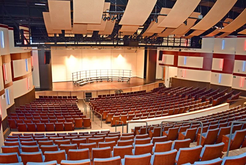 Inside an auditorium with a stage, rows of red seats, and decorative wall panels, ready for a performance or event.