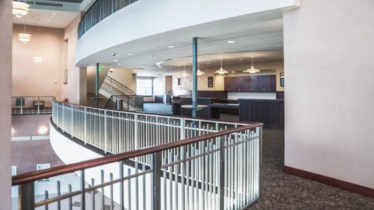 Interior of an empty multi-level building with a curved railing, carpeted floors, and a kitchenette area with dark wood cabinets and hanging lights.