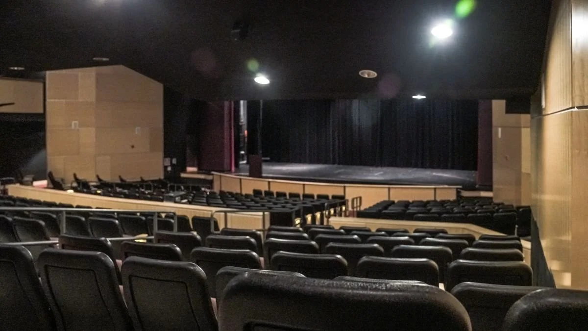 Empty theater with rows of black seats facing a stage with black curtains.