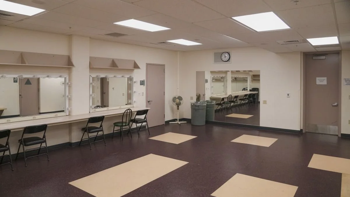 Empty dressing room with vanity mirrors, chairs, and bright ceiling lights.