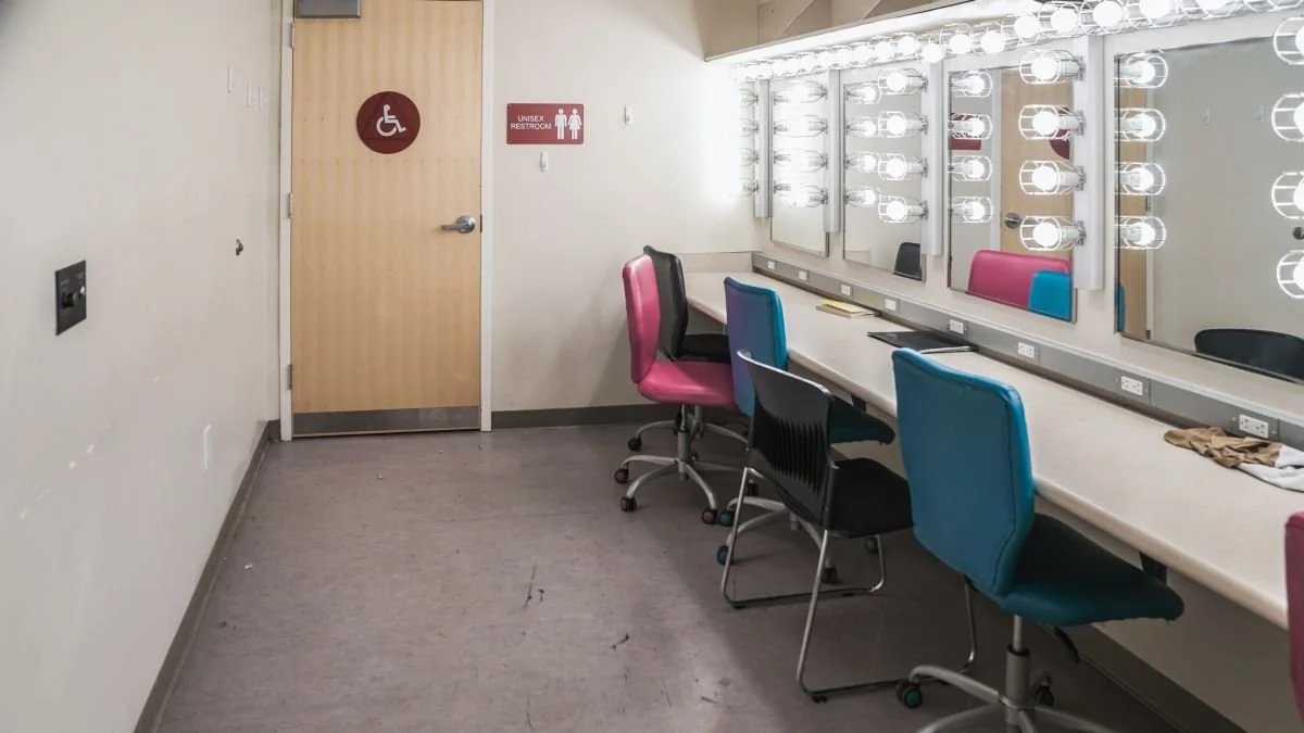Empty dressing room with makeup mirrors and chairs, equipped with bright lights, and a unisex restroom sign on the door.