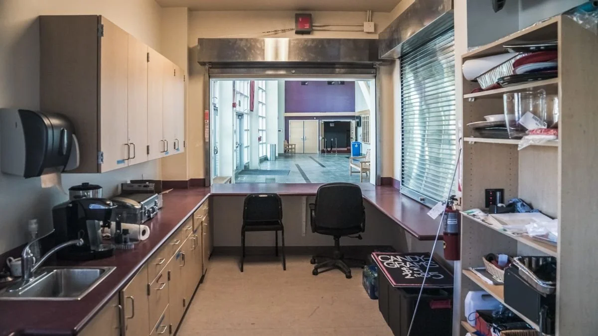 Kitchen area with cabinets, sink, coffee machine, and countertop facing an open window looking into a larger lobby or common area with chairs and a purple wall.