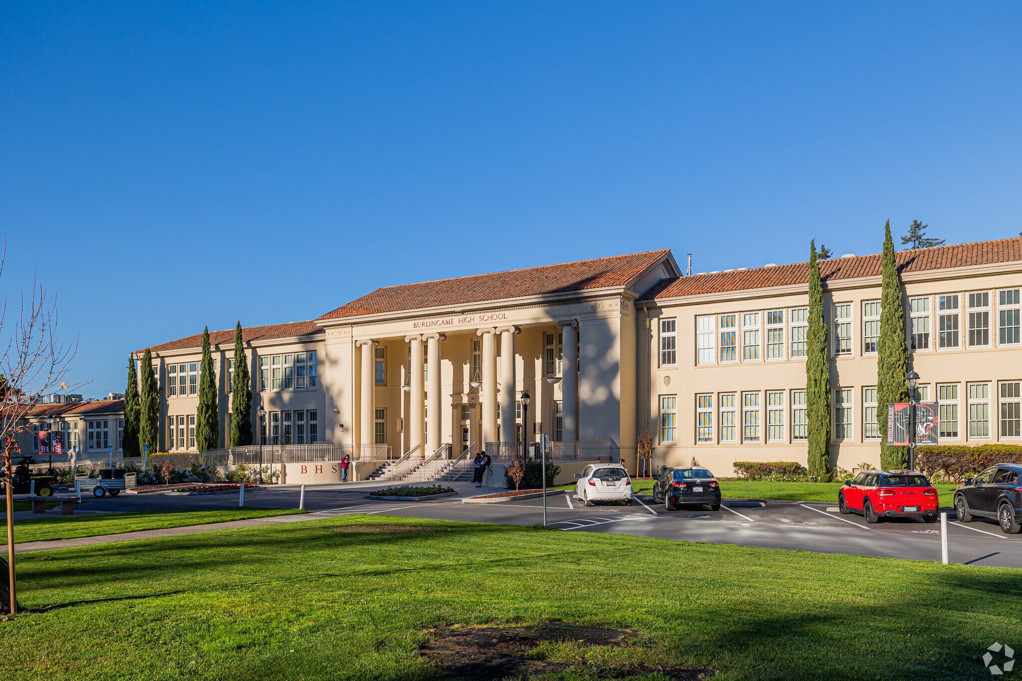 The exterior of Burlingame High School with a classical architectural style featuring cream-colored walls, tall columns, and a red-tiled roof. There are parked cars in front and a well-maintained green lawn.