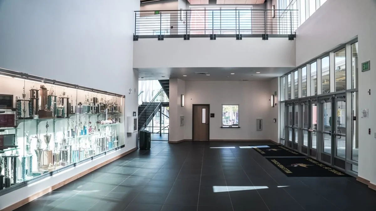 Empty lobby with a display case of trophies, glass double doors, and a staircase in the background.