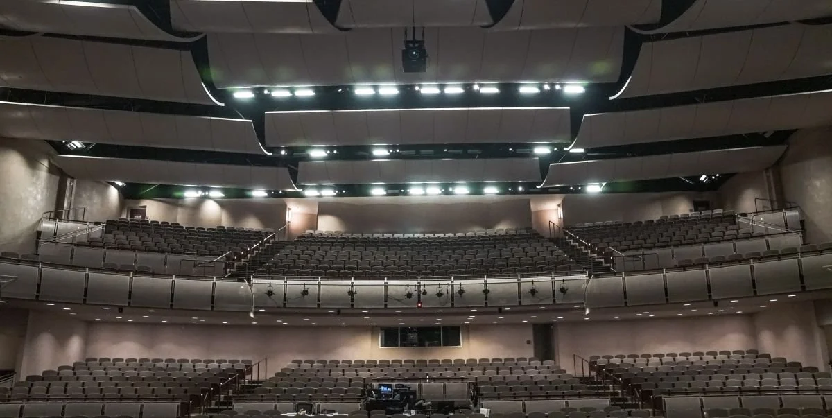 Empty theater auditorium with multiple tiers of seating, spotlighting, and a stage at the front.