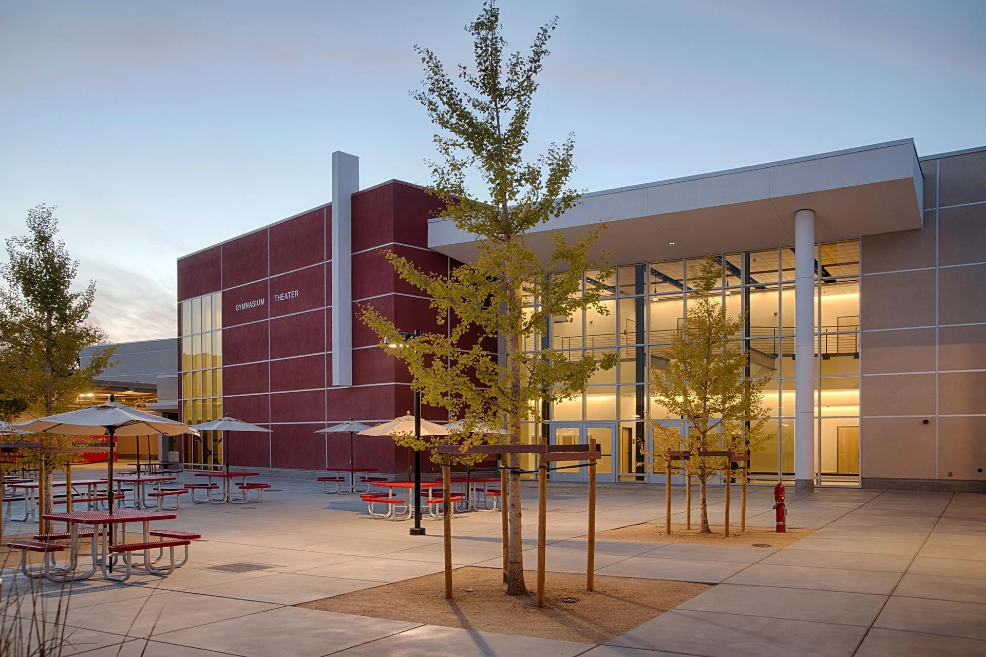 Modern school building with large glass windows, red accent wall labeled 'gymnasium' and 'theater', outdoor seating area with red picnic tables and umbrellas, young trees planted in the courtyard, during early evening.