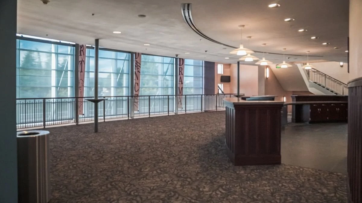 Empty indoor space with large windows, patterned carpet, wooden railings, and hanging lights.