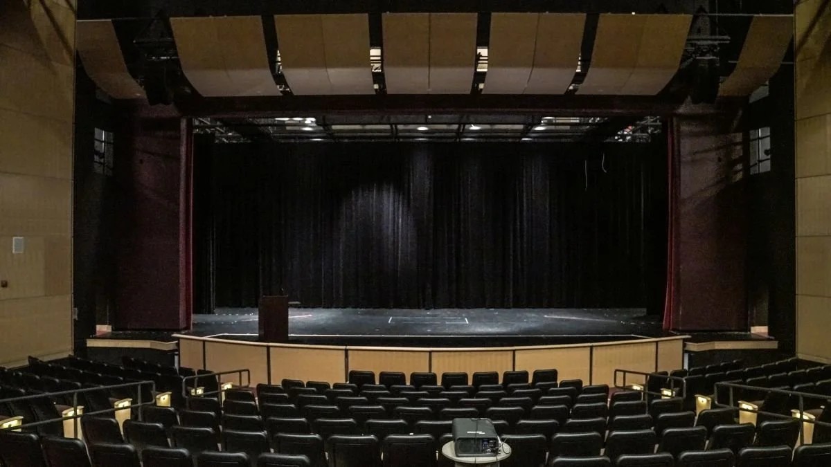 Empty theater stage with black curtains, surrounded by tiered seating, and a small table with audio equipment in front.