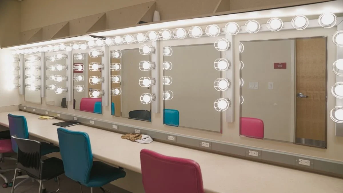 A row of four mirrors with bright Hollywood-style lights around the edges, mounted above a long countertop with colorful chairs and electrical outlets.
