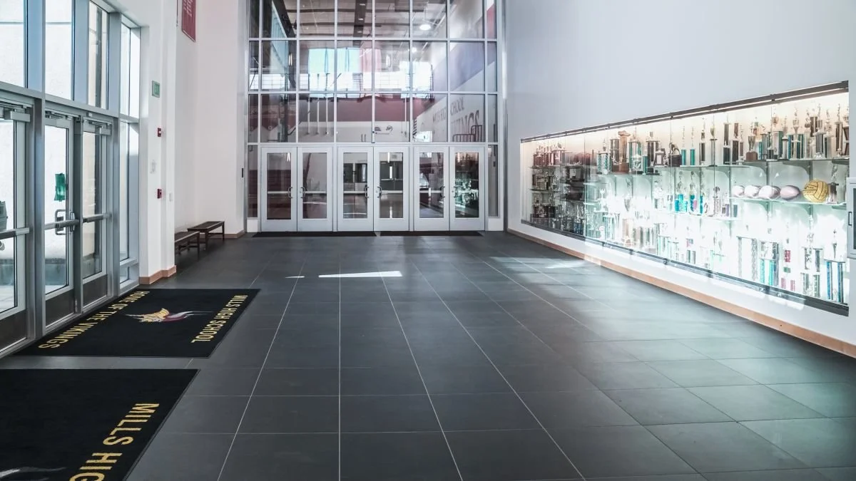 School lobby with glass doors, trophy display case, and black mats with yellow text at entrance.