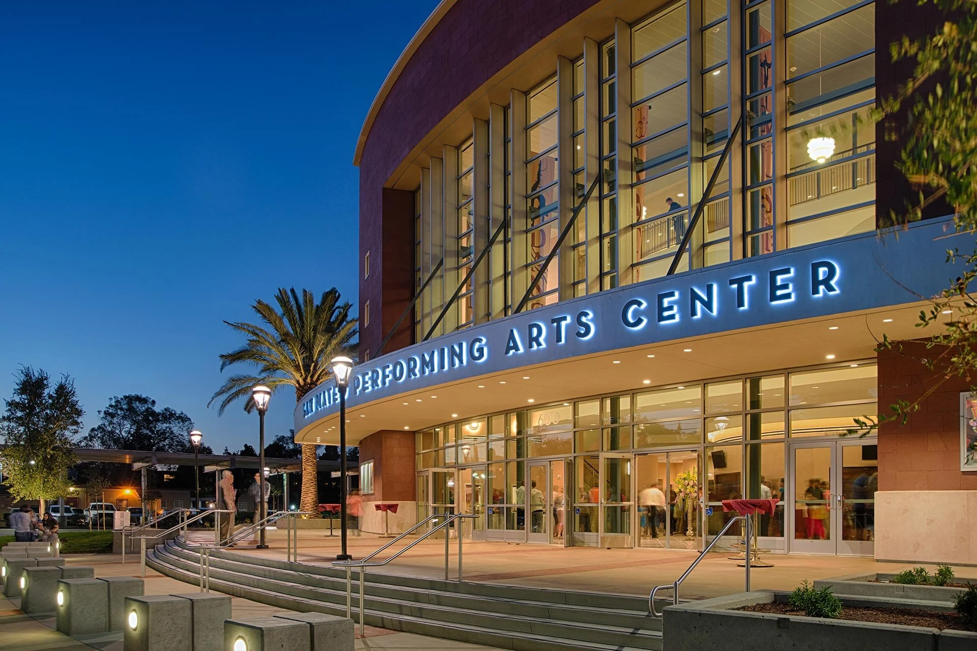 Exterior view of a performing arts center at dusk, featuring a curved glass facade with people inside, palm trees, street lamps, and a sign reading 'Performing Arts Center' with illuminated letters.