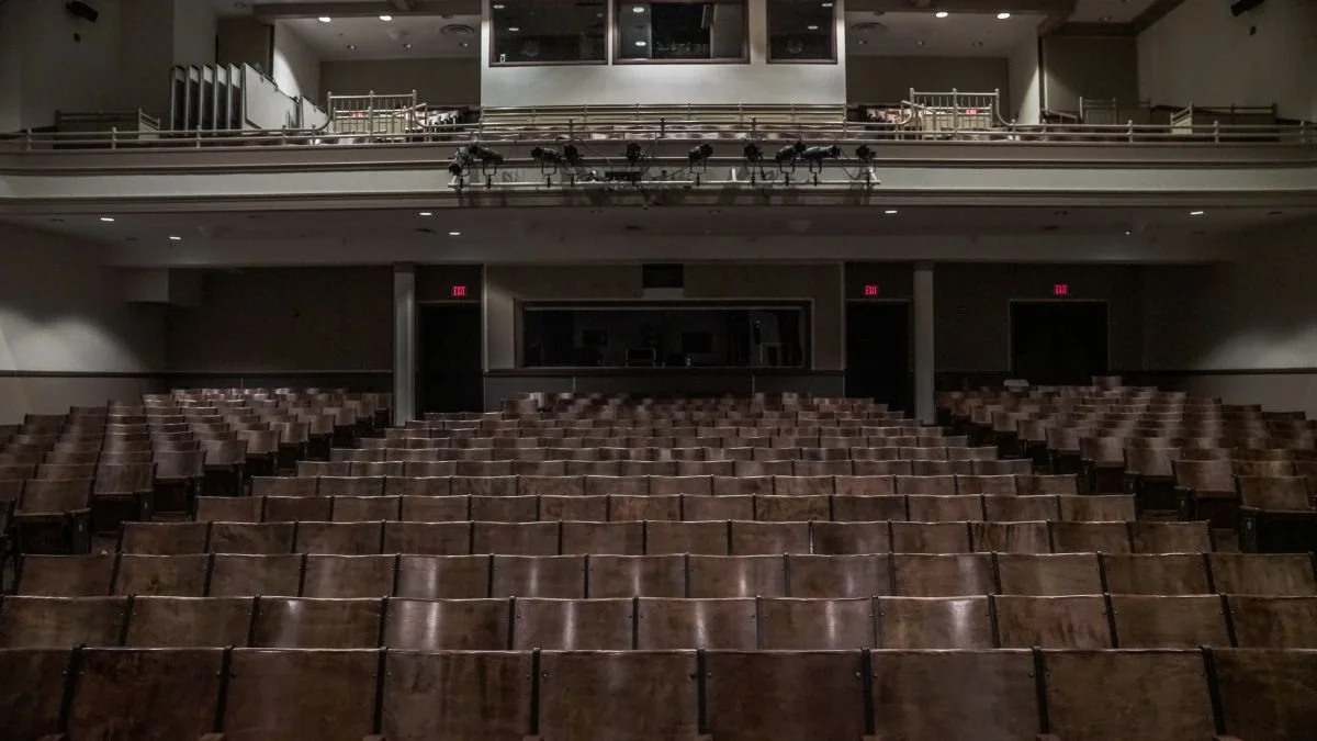 Empty theater or auditorium with rows of brown seats and a balcony level above.