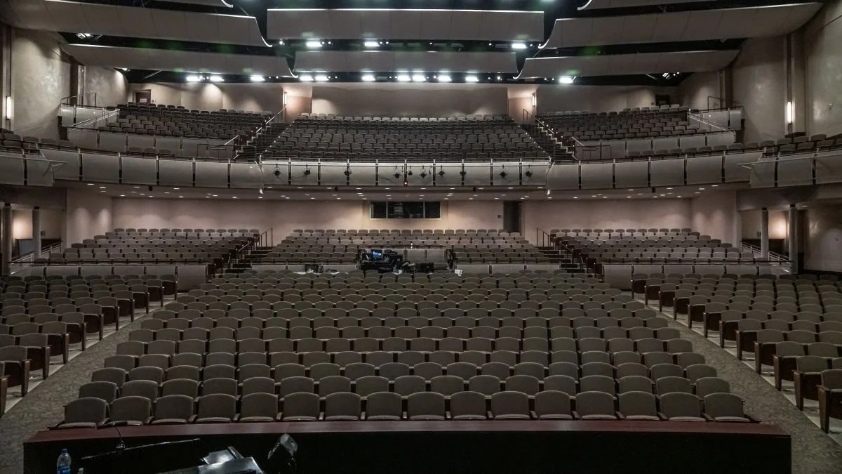 Empty theater auditorium with rows of seats, a stage, and lighting overhead.
