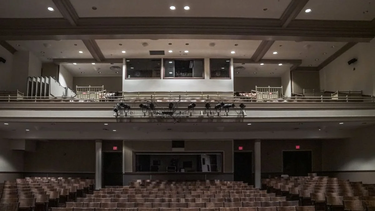 Empty theater auditorium with rows of chairs, a stage, and balcony seating, illuminated by overhead lighting.