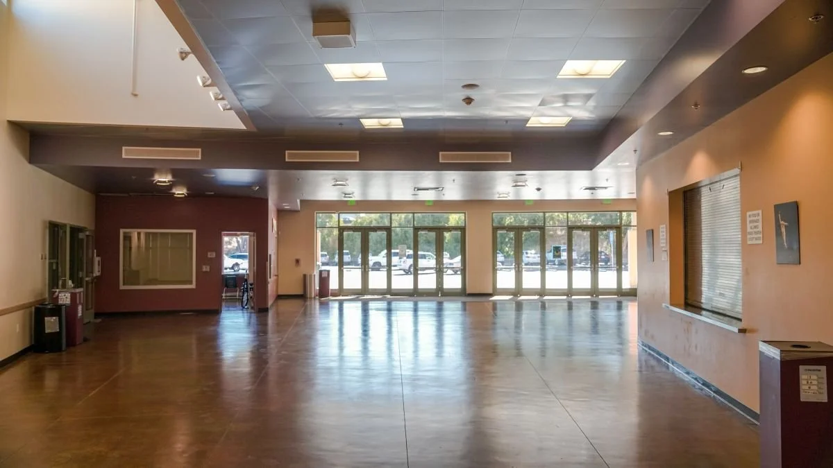 Empty large lobby or waiting area with wooden floors, glass doors, and some signage on the walls.