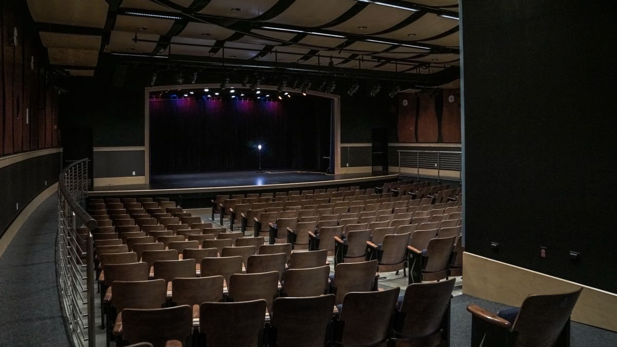 Empty theater with rows of brown seats facing a stage with black curtains and purple lighting.