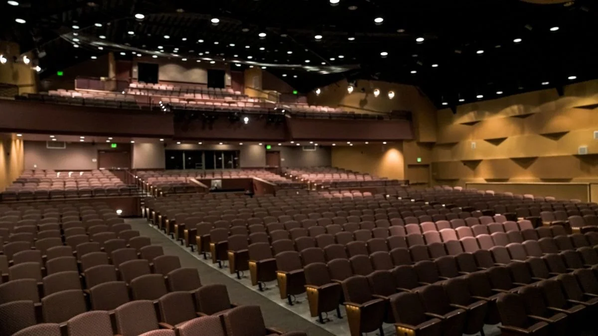 Empty theater with multiple rows of seats, a stage area, and ceiling lights.