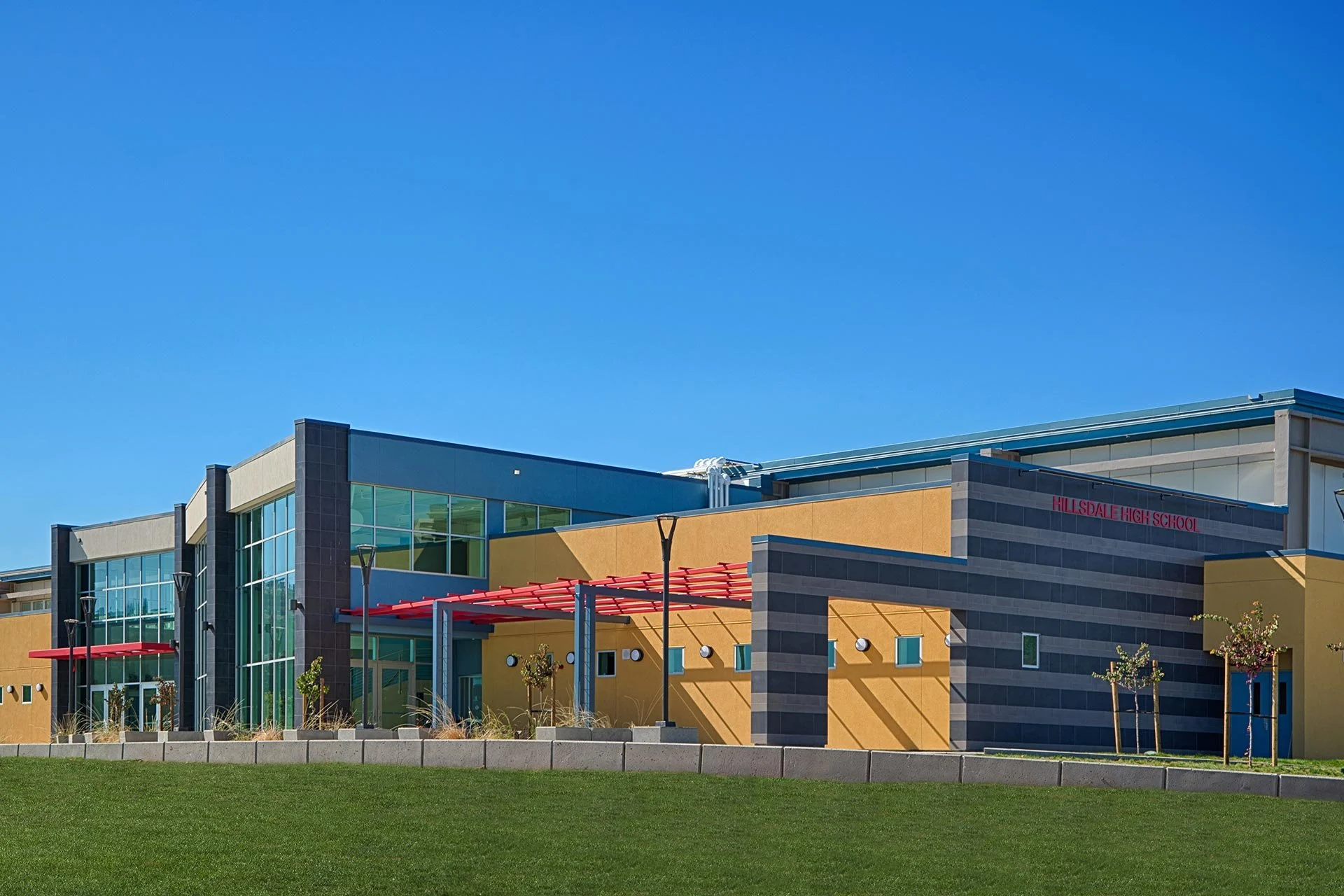Modern school building with yellow and gray exterior walls, large glass windows, and red accents, surrounded by a green lawn and small trees under a clear blue sky.