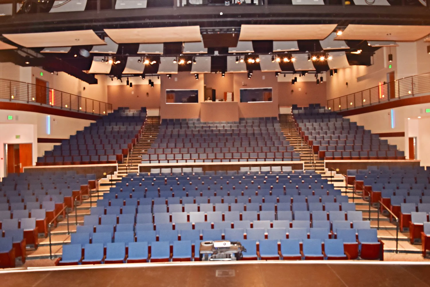 Empty auditorium with blue seats, stage at the front, and balcony level with rows of seats, ambient lighting, and ceiling acoustic panels.
