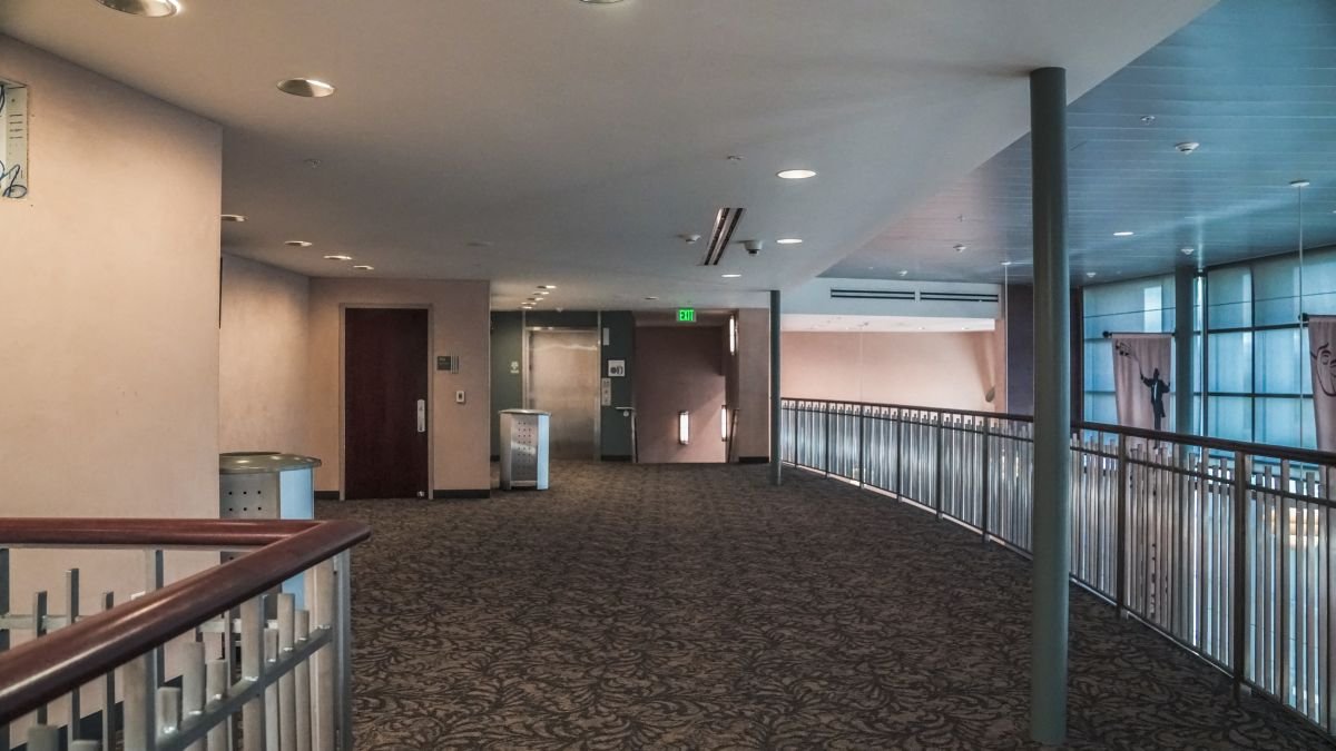 Empty hallway of a modern building with carpeted floor, beige walls, and several doors, with a railing on the right side and windows allowing natural light.