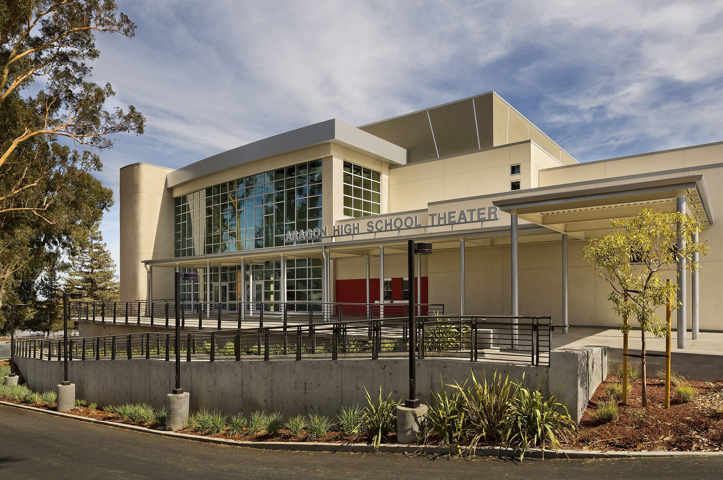 Exterior view of Aragon High School Theater building with large glass windows, a small tree in the foreground, and a clear blue sky.