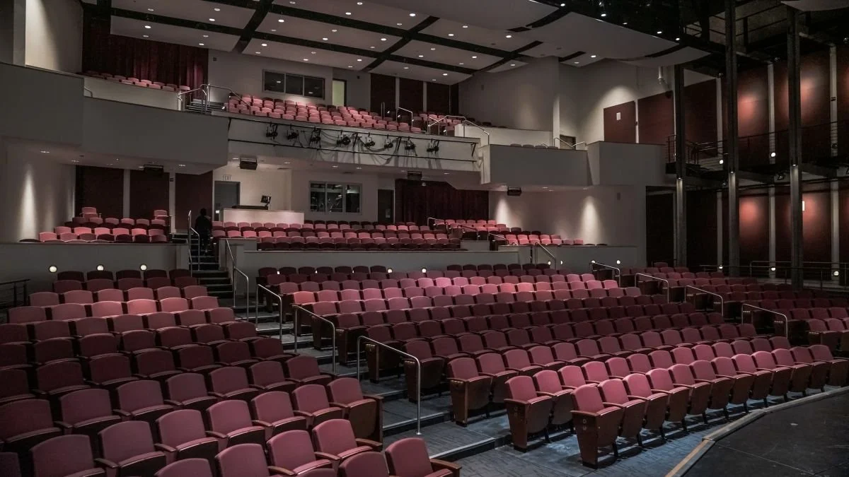 Interior of a theater or auditorium with rows of empty red seats and a stage or screen at the front.