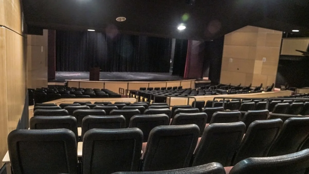 Empty theater with rows of black seats facing a stage with closed black curtains.