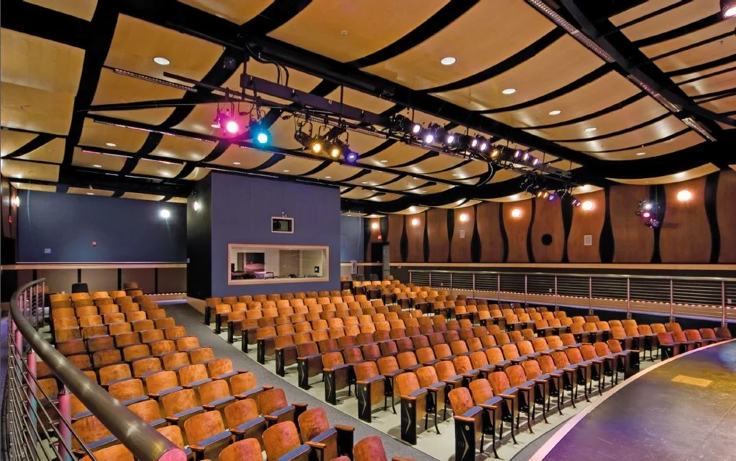Empty theater with rows of orange seats, stage on the right, black acoustic panels on the walls, and stage lighting overhead.