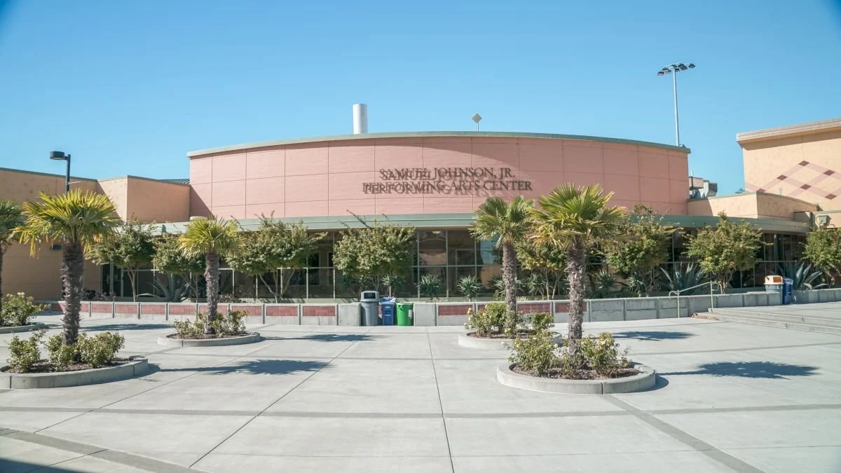 Exterior view of the Samuel Johnson Jr. Performing Arts Center with palm trees and trash bins in front, under a clear blue sky.