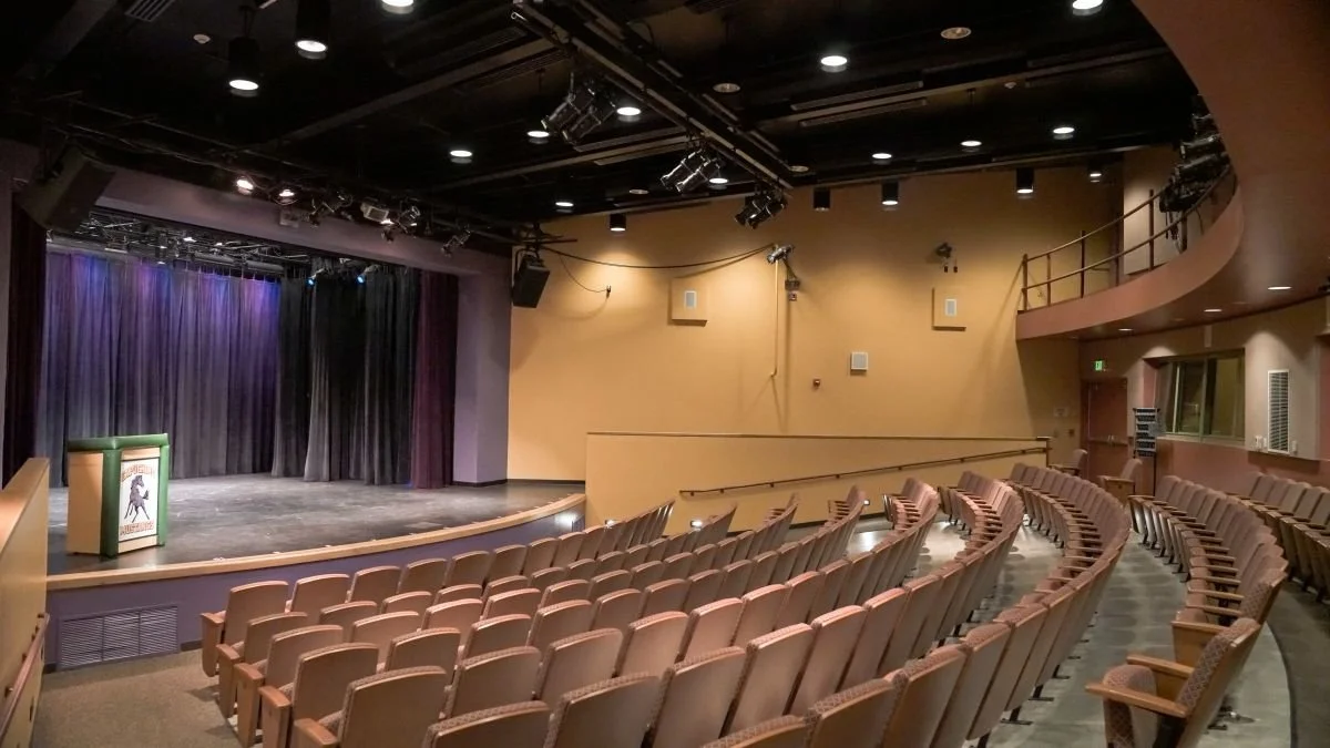 Empty auditorium with a stage, curtains, and beige chairs arranged in rows.